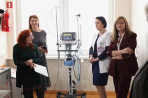 W sali medycznej cztery kobiety stoją obok nowoczesnego respiratora Hamilton Medical. Od lewej: kobieta z rudymi włosami (Renata Polisiakiewicz), trzyma teczkę i kulę, kobieta w ciemnej sukience w kropki, lekarka (Renata Nieznaj) w białym fartuchu oraz kobieta w bordowej marynarce (Ewa Mańdziuk). Na ścianie po lewej widoczna czerwona gaśnica