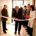 Uroczyste otwarcie wypożyczalni sprzętu rehabilitacyjnego przy SION w Bartoszycach. Pięć osób stoi przed biało-czerwoną wstęgą, jeden z mężczyzn przecina ją nożyczkami, w tle widoczne inne osoby przy wejściu