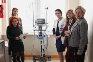 Pięć kobiet pozuje do zdjęcia. Wszystkie są zwrócone w stronę obiektywu i uśmiechają się. Po lewej stronie stoją dwie kobiety (jedna z rudymi włosami i kulą), po prawej trzy pozostałe, w tym kobieta w szarym żakiecie. W centrum stoi respirator medyczny Hamilton Medical. Pomieszczenie jest bardzo jasne, oświetlone światłem wpadającym przez duże okno w tle