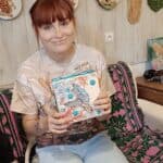 Uśmiechnięta młoda dziewczyna siedzi oparta o fotel z wzorzystą narzutą. Prezentuje skrzynkę z decoupage przedstawiającym żółwia morskiego. W tle na ścianie prace plastyczne na plastrach drewna i wyciskane z roślin