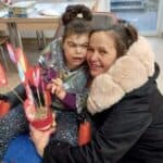 Małgorzata w czarnej odzieży z jasnym futrzanym kołnierzem przytula policzek do policzka córki Urszuli. W rękach trzyma doniczkę z papierowymi kwiatami i sercami na drewnianych patyczkach