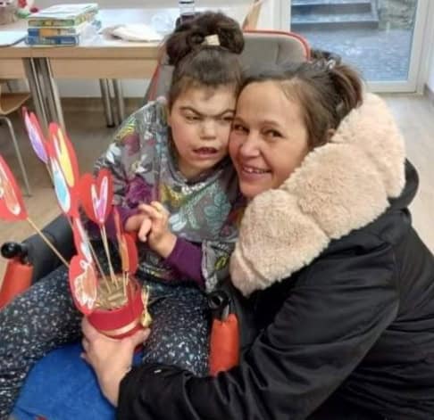 Małgorzata w czarnej odzieży z jasnym futrzanym kołnierzem przytula policzek do policzka córki Urszuli. W rękach trzyma doniczkę z papierowymi kwiatami i sercami na drewnianych patyczkach