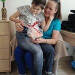 Na kolanach uśmiechniętej Małgorzaty Szestopałko siedzi córka Urszula. Kobiety patrzą na przedmiot zapakowany w celofan i przewiązany czerwoną wstążką