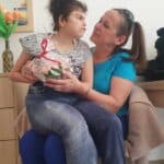 Na kolanach Małgorzaty Szestopałko siedzi, obrócona bokiem, córka Urszula. Kobiety patrzą na siebie. Jedna z nich trzyma przedmiot zapakowany w celofan i przewiązany wstążką