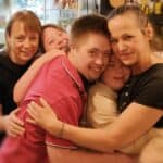 Pięć osób przytula się do siebie i uśmiecha. Z przodu Małgorzata Szestopałko obejmuje młodą kobietę i mężczyznę, za nimi stoją dwie kobiety. W tle przy oknie witrynki meblarskie z ekspozycją prac