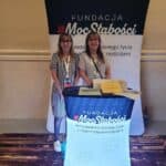 Magdalena w sukience w biało-czarne pionowe paski oraz Marta w beżowej sukience stoją na tle rollupa z nazwą Fundacji Moc Słabości. Przed nimi pulpit z nazwą Fundacji oraz zwrotem "Ambasadorki dobrego życia z niepełnosprawnościami"