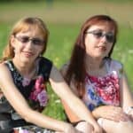 Siostry Wróbel siedzą na zielonej łące. Obie kobiety mają długie włosy i grzywki, ubrane są w sukienki w kwiaty