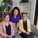 Siostry Wróbel siedzą na szarej kanapie, za która stoi Anna Dymna. Marta z lewej strony ubrana w granatową sukienkę i żakiet w kolorze pudrowy róż. Magda w małej czarnej z koronkowym dekoltem oraz żółtej marynarce