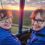 Uśmiechnięte Magda i Marta ubrane w ciemnoniebieskie wiatrówki stoją w koszu balonu unoszącego się w powietrzu ponad zielonymi polami i lasami