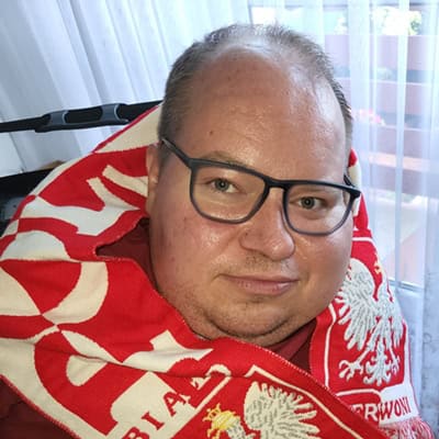 Paweł G. Iwaniuk w okularach patrzy prosto w obiektyw. Ma na szyi biało-czerwony szalik kibica Polski z godłem państwa.