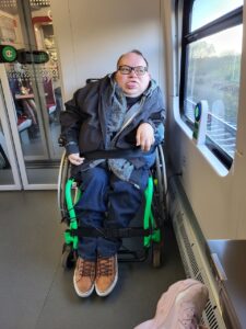 Wnętrze pociągu z oknem oraz przeszklonymi drzwiami do przedziału w tle. Przy oknie siedzi Paweł G. Iwaniuk, mężczyzna w okularach przeciwsłonecznych, ubrany w bluzę z kapturem i kurtkę. Mężczyzna obserwuje przez okno mijany krajobraz.