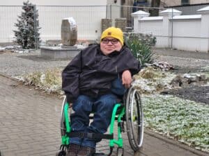 Paweł G. Iwaniuk, mężczyzna w okularach, na wózku, ubrany w czarną kurtkę i żółtą czapkę, pozuje na chodniku w plenerze obok trawnika opruszonego śniegiem. W tle na cokole kamień z wykutym napisem „ODRODZENIE OJCZYZNY 1918”.