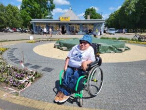 Paweł G. Iwaniuk, mężczyzna w okularach, na wózku, ma T-shirt i kapelusz typu bucket hat, pozuje na tle parterowego pawilonu w Jeruzalu o błękitnej elewacji i witrynach z żółto-granatowymi napisami, szerzej znanego z serialu „Ranczo” jako sklep w Wilkowyjach. Na pawilonie szyld „U KRYSI. SKLEP SPOŻYWCZO_PRZEMYSŁOWY Wł. Krystyna Węcławska”.