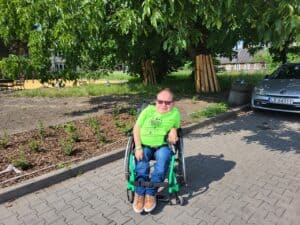 Paweł G. Iwaniuk pozuje w plenerze przy słonecznej pogodzie, stoi pod drzewem z bujnymi zielonymi liśćmi. Mężczyzna jest ubrany w zielony T-shirt z napisem „Mistrzowie kręcą dla Białej Podlaskiej”. W tle zabudowania.