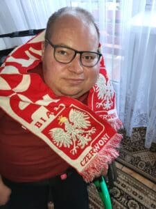 Portret Pawła G. Iwaniuka w okularach. Ma na szyi biało-czerwony szalik kibica Polski z godłem państwa. Mężczyzna patrzy prosto w obiektyw, stoi przy oknie z długą, białą firanką sięgającą podłogi, na której leży wzorzysty dywan.