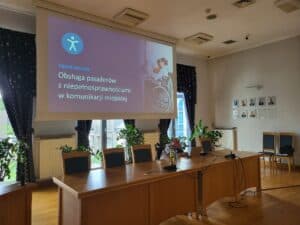 W przestrzeni konferencyjnej przy oknie z długim, sześcioosobowym stołem siedzi Paweł G. Iwaniuk. Mężczyzna patrzy w rozłożony laptop, który stoi przed nim. Od sufitu zwisa ekran multimedialny z wyświetlonym slajdem: „Paweł Iwaniuk: Obsługa pasażerów z niepełnosprawnościami w komunikacji miejskiej”.