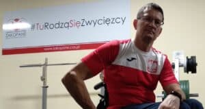 Zgrupowanie Kadry Narodowej Osób Niepełnosprawnych w łucznictwie, Zakopane 2020. Piotr Jacniacki siedzi na wózku. Ma na sobie biało-czerwoną koszulkę sportową z godłem Polski na piersi. W tle widoczna jest sztanga oraz jasna ściana z logotypem „Centralny Ośrodek Sportu Zakopane” i hasłem „#TuRodząSięZwycięzcy”