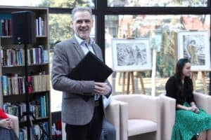 Janusz Padziński uśmiecha się szeroko do obiektywu. Ubrany w szarą, melanżową marynarkę i wzorzystą koszulę. Oburącz trzyma przed sobą elegancką, czarną aktówkę. Stoi w jasnym pomieszczeniu obok regału wypełnionego książkami i czarnego głośnika na statywie. W tle, za dużym przeszkleniem, widać drzewa iglaste, a wewnątrz sali sztalugi z czarno-białymi grafikami przedstawiającymi anioły i sceny figuralne.