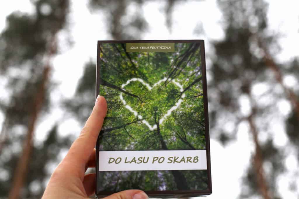 Dłoń trzyma prostokątne pudełko gry terapeutycznej „Do lasu po skarb” na tle rozmytego lasu iglastego. Na okładce gry widnieje zdjęcie koron drzew widzianych z dołu, w których prześwit nieba układa się w kształt serca. W górnej części znajduje się napis „GRA TERAPEUTYCZNA”, a w dolnej, na białym pasku, tytuł „DO LASU PO SKARB”.