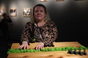 Portret kobiety o jasnych włosach, stojącej przy drewnianym blacie z pasem zielonego mchu. Kobieta dotyka mchu obiema dłońmi, lekko się uśmiechając. Jest ubrana w ciemną bluzkę w beżowe wzory. W tle widać rozmyte sylwetki innych osób i prace na ścianach