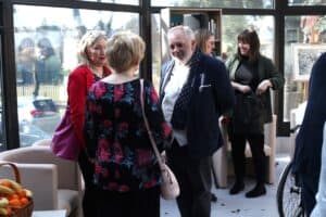 Grupa uczestników rozmawia w kuluarach. W centrum stoi starszy mężczyzna w ciemnej marynarce i wzorzystym szalu, uśmiecha się do kobiety w czarnej bluzce w kwiaty. Obok stoi kobieta w czerwonym żakiecie z fioletową torebką. W tle widać inne rozmawiające osoby oraz regał z książkami.