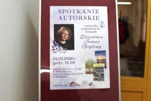 Pionowy plakat informacyjny umieszczony w srebrnej ramie. Na białym tle duży fioletowy napis „SPOTKANIE AUTORSKIE”. Poniżej zdjęcie portretowe dojrzałej, uśmiechniętej blondynki w okularach (Zdzisława Joanna Gigling) oraz grafika z fioletowym motylem i kwiatami. Po prawej stronie widnieje ozdobny napis z imieniem i nazwiskiem autorki. Na dole plakatu data 04.03.2026 r., godzina 11.00 oraz adres: Galeria Kultury „Krąg” w Lublinie. Widoczne są również miniatury okładek trzech książek: „Księżyc nad jeziorem”, „Tajemnica starego kufra” oraz „Dwadzieścia pięć lat później”