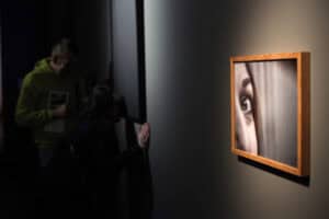 Zbliżenie na fotografię w drewnianej ramie, przedstawiającą detal twarzy z wyraźnie zarysowanym, szeroko otwartym okiem. W tle, w głębi sali, widać nieostre sylwetki dwojga zwiedzających: mężczyzny w zielonej bluzie oraz kobiety nachylonej przy czarnej ścianie. Kompozycja podkreśla temat patrzenia i obserwacji