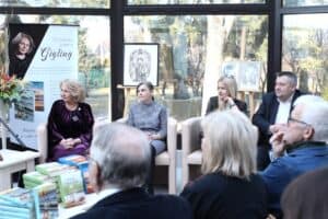 W nowoczesnej, przeszklonej sali siedzi czworo panelistów (Zdzisława Joanna Gigling, dr Magdalena Czarnecka, Anna Augustyniak i Grzegorz Sołtys) w beżowych, nowoczesnych fotelach. Po lewej stronie stoi wysoki roll-up promujący twórczość Zdzisławy Joanny Gigling. Na niskim stoliku na pierwszym planie ułożone są stosy książek w twardych oprawach. W tle, za oknami, rozpościera się widok na nasłoneczniony ogród, a wewnątrz sali ustawione są sztalugi z pracami artystycznymi