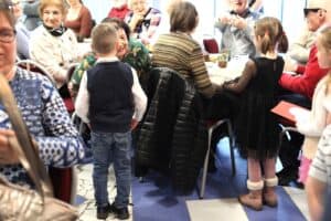 Dzieci wręczają kobietom siedzącym przy stołach, laurki i kartki z okazji Dnia Kobiet