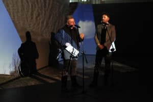 Dwóch mężczyzn stoi przy mikrofonach na tle wielkoformatowych projekcji wideo. Mężczyzna po lewej (Marek Lapis), w niebiesko-czarnej marynarce, trzyma w ręku folder i gestykuluje. Mężczyzna po prawej, w jasnej marynarce, słucha z rękami splecionymi przed sobą. Na ścianach za nimi widać fragmenty krajobrazu z błękitnym niebem i chmurami