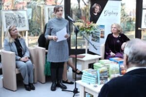dr Magdalena Czarnecka w sukience w biało-czarną „pepitkę” stoi przy mikrofonie i czyta tekst z białej kartki A4. W tle siedzi autorka, Zdzisława Joanna Gigling, spogląda w jej stronę. Obok siedzi inna kobieta w szarym żakiecie. Na stoliku przed nimi piętrzą się egzemplarze książek.