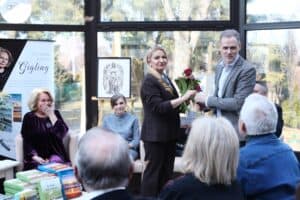 Janusz Padziński wręcza kwiaty Annie Augustyniak. W tle Zdzisława Joanna Gigling reaguje spontanicznym uśmiechem, zakrywa dłońmi usta. Widownia, widoczna z tyłu, obserwuje tę uroczystą chwilę. W tle na sztaludze widnieje grafika przedstawiająca anioła