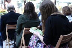 Perspektywa zza pleców publiczności. Na pierwszym planie kobieta o długich włosach siedząca na drewnianym krześle trzyma w dłoniach książkę z zieloną okładką. Przygląda się zdjęciu autorki i biogramowi na skrzydełku oprawy. W tle widać nieostre sylwetki innych osób oraz fragment dekoracji sali