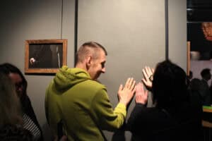 Uśmiechnięty mężczyzna w zielonej bluzie z kapturem oraz kobieta w ciemnym ubraniu dotykają otwartymi dłońmi dużej, szarej płaszczyzny pracy artystycznej w ramie. Oboje są zwróceni do siebie profilami. Po lewej stronie, na ścianie, wisi mniejsza fotografia przedstawiająca dłonie i białą laskę