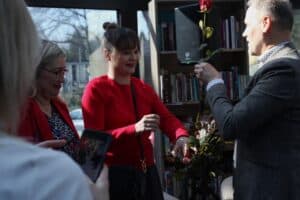 Janusz Padziński podaje pojedynczą czerwoną różę o długiej łodydze kobiecie ubranej w intensywnie czerwoną bluzkę. Kobieta uśmiecha się promiennie. W tle widać fragment regału bibliotecznego oraz czarny głośnik
