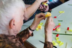 Dłonie seniorki w brązowej bluzce, która za pomocą cienkiej nitki zabezpiecza płatki sztucznego kwiatu. Na stole obok niej leżą gotowe żółte żonkile oraz płatki wycięte z papieru