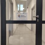 Przeszklone drzwi w brązowej ramie; na drzwiach pozioma kartka z napisem ‚Regionalne Centrum Komunikacji, ul. Do Dysa 5, piętro I, 20-149 Lublin’ oraz logotypami unijnymi i partnerskimi. W tle widać długi, jasno oświetlony korytarz biurowy z wieloma drzwiami