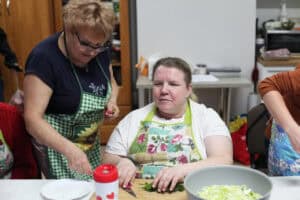 Kobieta w białym t-shircie i kwiecistym fartuchu sieka koperek na desce, obok druga kobieta udziela wskazówek