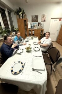 Widok jadalni – trzy kobiety siedzą przy nakrytym stole i czekają na posiłek. Na stole rozstawione są talerze z niebiesko - żółtą obwódką, sztućce oraz szklana miska z zieloną sałatką