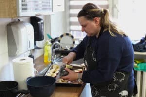 Kobieta o jasnych włosach upiętych w kucyk, ubrana w ciemny fartuch z białym nadrukiem owieczek, wsypuje rodzynki do wydrążonych połówek jabłek ułożonych na blasze