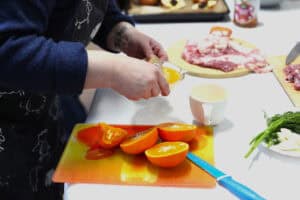 Dłonie osoby w fartuchu w owieczki wyciskają sok z pomarańczy na ręcznej wyciskarce; obok leżą czosnek i koperek