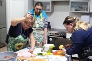Kobieta w okularach i zielonym fartuchu w słoneczniki zszywa kaczkę nicią kuchenną; obok druga osoba obiera cytrynę. W tle stoi uczestniczka, obserwuje postępy