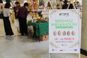 Pionowy biały potykacz reklamowy z logo "ROPS Lublin" i napisem: „KIERMASZ WIELKANOCNY Podmiotów Ekonomii Społecznej 28 - 29 MARCA, Galeria Olimp w Lublinie"; w tle stoiska
