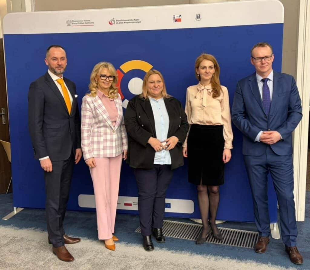 Pięć osób – między dwoma mężczyznami stoją trzy kobiety – pozuje do zdjęcia na tle ścianki z logotypem Centrum Komunikacji dla Osób z Niepełnosprawnościami