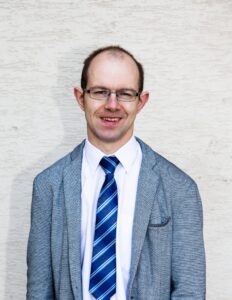 Uśmiechnięty mężczyzna w średnim wieku, Krótkie włosy, metalowe, prostokątne okulary. Ubrany w białą koszulę, krawat w granatowe, niebieskie i białe paski oraz szaro-niebieską marynarkę