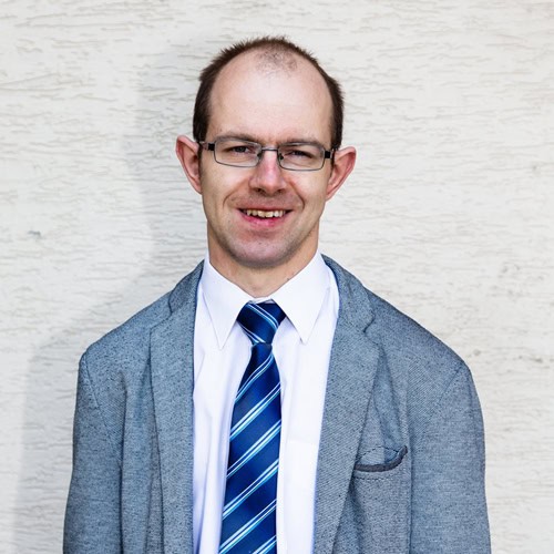 Portret Krzysztofa Kurowskiego. Uśmiechnięty mężczyzna w średnim wieku, Krótkie włosy, metalowe, prostokątne okulary. Ubrany w białą koszulę, krawat w granatowe, niebieskie i białe paski oraz szaro-niebieską marynarkę