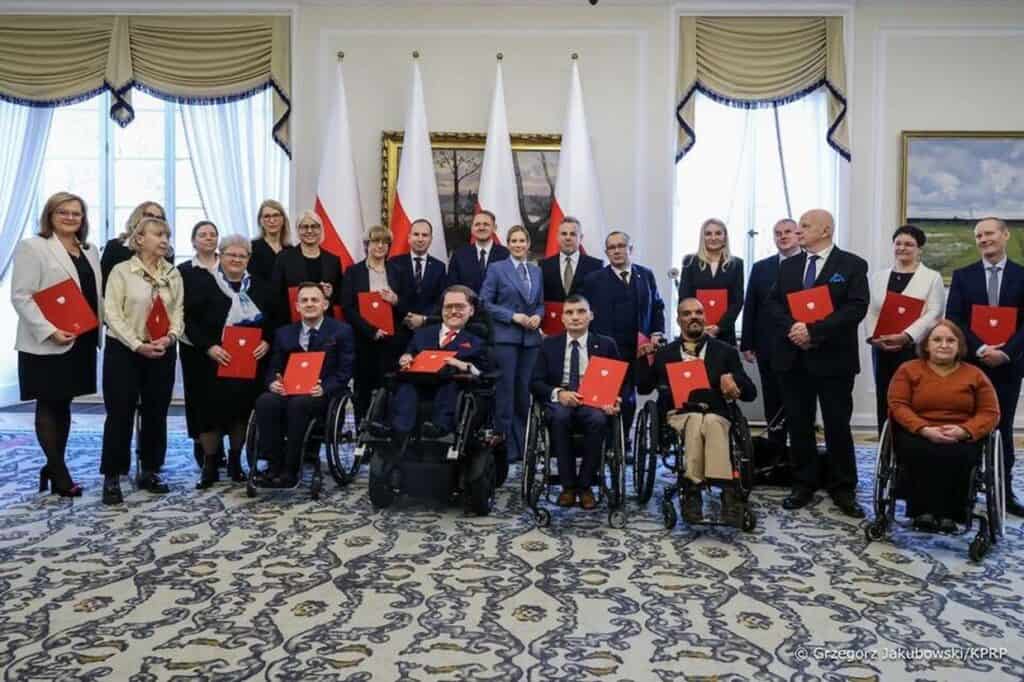 Grupa osób (członków nowo powołanej Rady do spraw Osób z Niepełnosprawnościami) pozuje do wspólnego zdjęcia w sali Pałacu Prezydenckiego. Część osób siedzi na wózkach inwalidzkich, pozostali stoją. Większość trzyma czerwone teczki z godłem Polski. W tle widoczne są flagi państwowe.