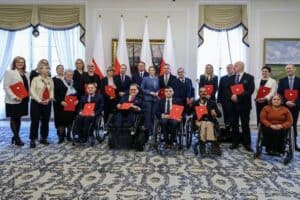 Grupa osób (członków nowo powołanej Rady do spraw Osób z Niepełnosprawnościami) pozuje do wspólnego zdjęcia w sali Pałacu Prezydenckiego. Część osób siedzi na wózkach inwalidzkich, pozostali stoją. Większość trzyma czerwone teczki z godłem Polski. W tle widoczne są flagi państwowe.