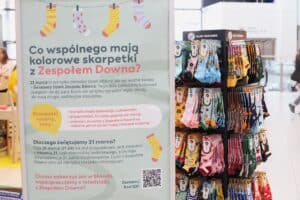 Pionowa tablica informacyjna w pasażu galerii handlowej pt. „Co wspólnego mają kolorowe skarpetki z Zespołem Downa?, uzupełniony rysunkami kolorowych skarpetek oraz kodem QR. Obok obrotowy stojak z ekspozycją skarpetek marki Many Mornings