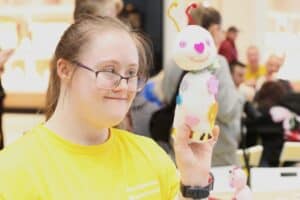 Wolontariuszka w okularach i żółtej koszulce spogląda z uśmiechem na trzymaną w dłoni maskotkę wykonaną z białej skarpetki z różowymi oczami, nosem w kształcie serca, uśmiechniętą buzię oraz długimi, brokatowymi czułkami z czerwonych i żółtych drucików kreatywnych